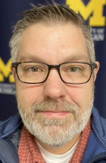 Color headshot of a white male looking at the camera. He has dark brown hair, is clean-shaven, and wearing a French blue, long-sleeve shirt